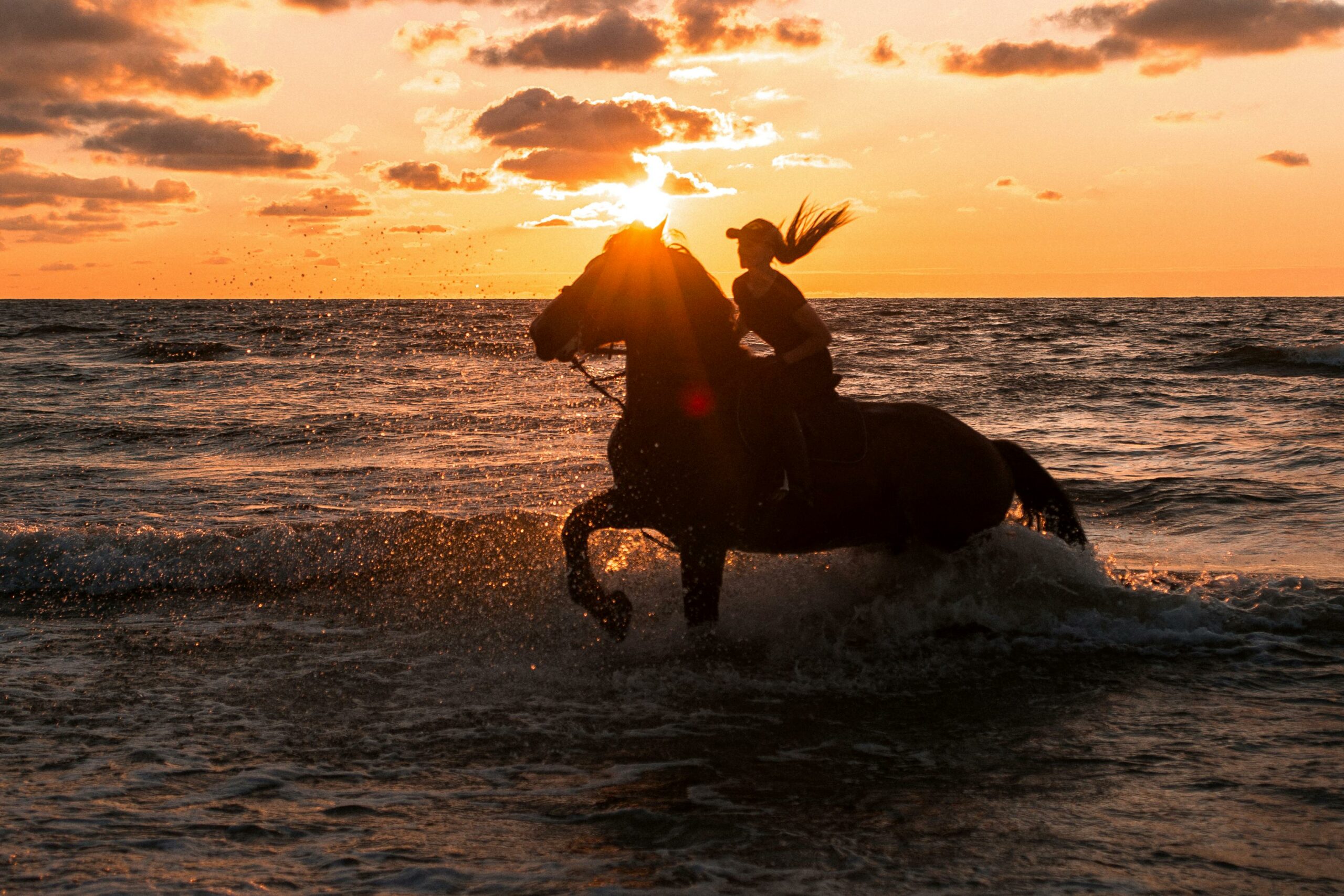 Silhouette of a woman riding a horse in the sea at sunset, creating a dramatic coastal scene.